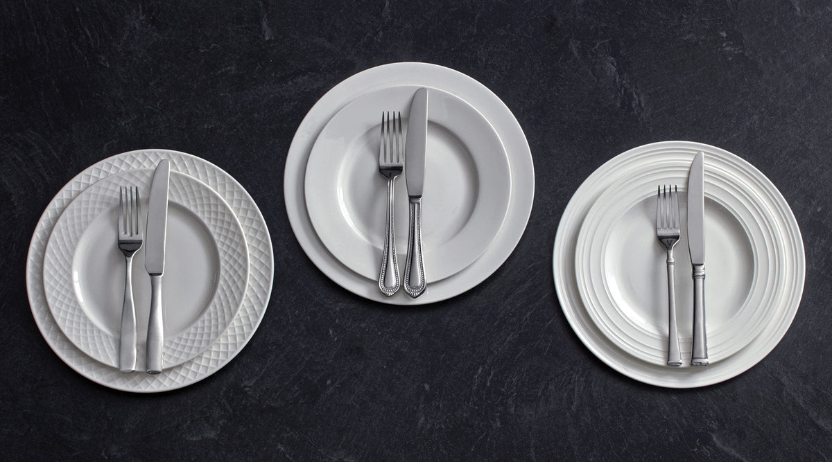 Three white dinnerware place settings on a dark surface with a textured plate stack on the left, plain plates in the center with silverware, and a ridged plate stack on the right.