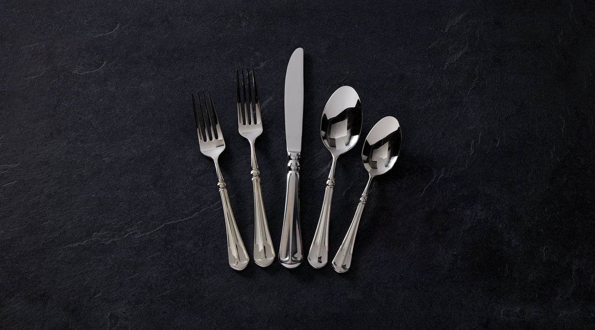 Six piece polished stainless steel flatware arranged in a fan on a dark slate surface including three forks, a knife, and two spoons