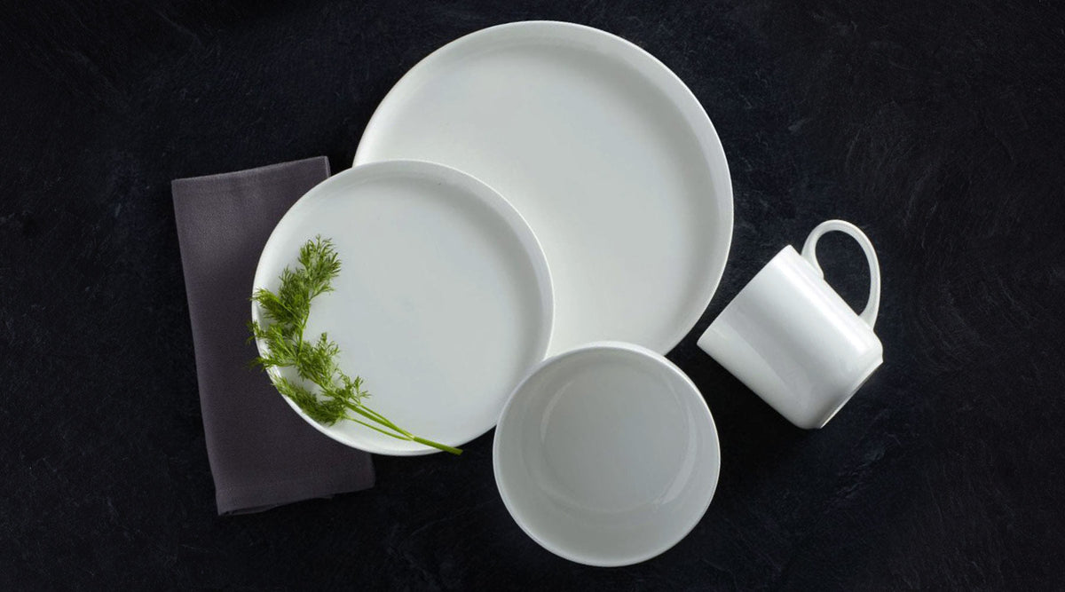 Samantha white ceramic dinnerware set with plates bowls and a mug arranged on a dark surface with dill sprigs.