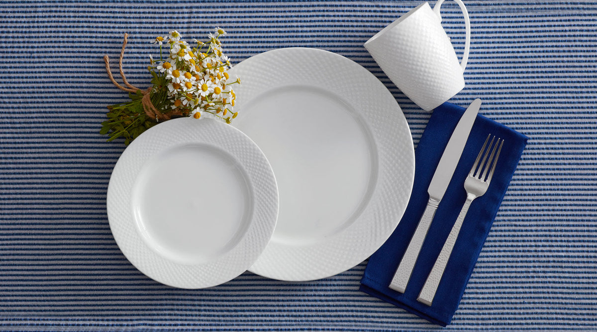Stanton table setting with stacked white textured plates, a white mug, a small bouquet of daisies, and a knife and fork on a blue napkin.