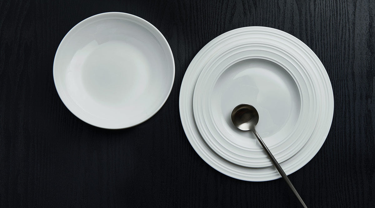White ceramic bowl beside a stack of white plates with a metal spoon resting in the bottom plate on a dark wood surface