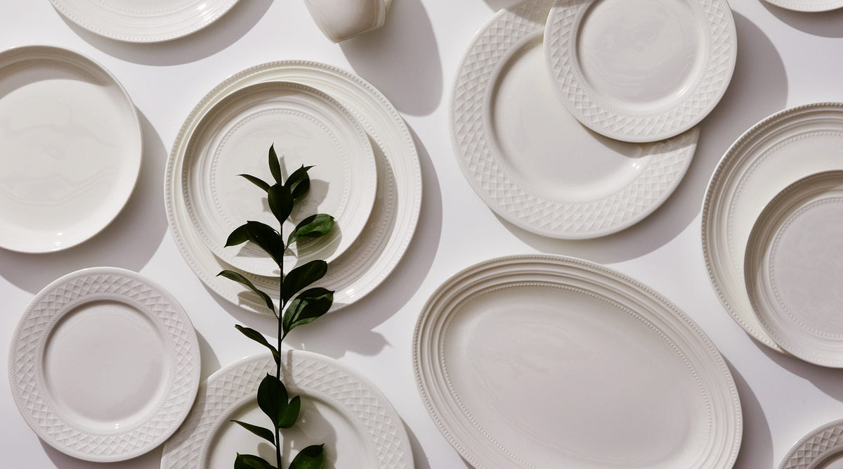 Flat lay of white Vegan Bone China plates with textured rims arranged on a white background, accented by a green leafy stem.