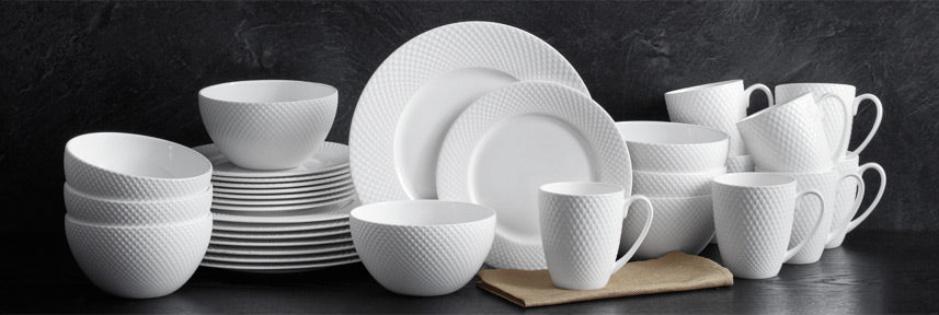 White bone china dinnerware including plates bowls and mugs with a textured dotted pattern arranged on a dark surface.
