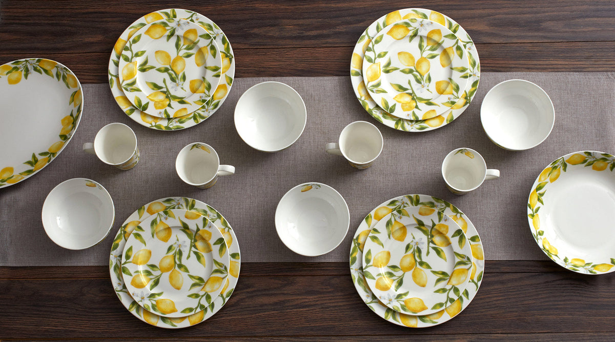 White porcelain plates with yellow lemon prints and matching mugs arranged on a dark wooden table with a gray table runner.