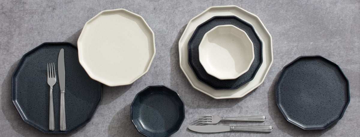 Luxor dinnerware collection arranged on a gray surface featuring navy speckled plates and ivory bowls with silverware.