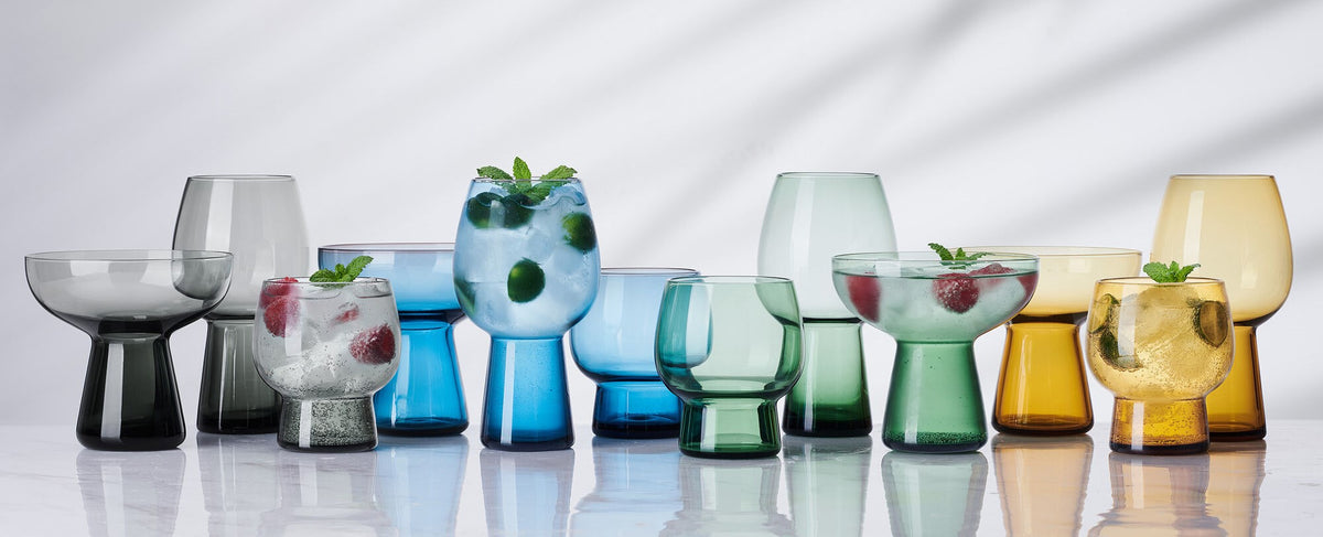 Phoebe glassware collection: row of colorful goblets, tumblers, and bowls in blue green and amber on a reflective surface, some with mint garnish.