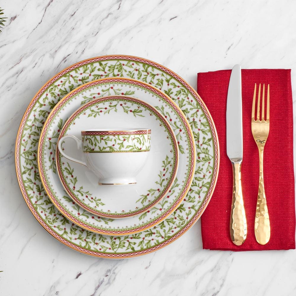 Holiday Traditions dinnerware set with holly patterned plates and a matching teacup on a marble table with a red napkin and gold utensils